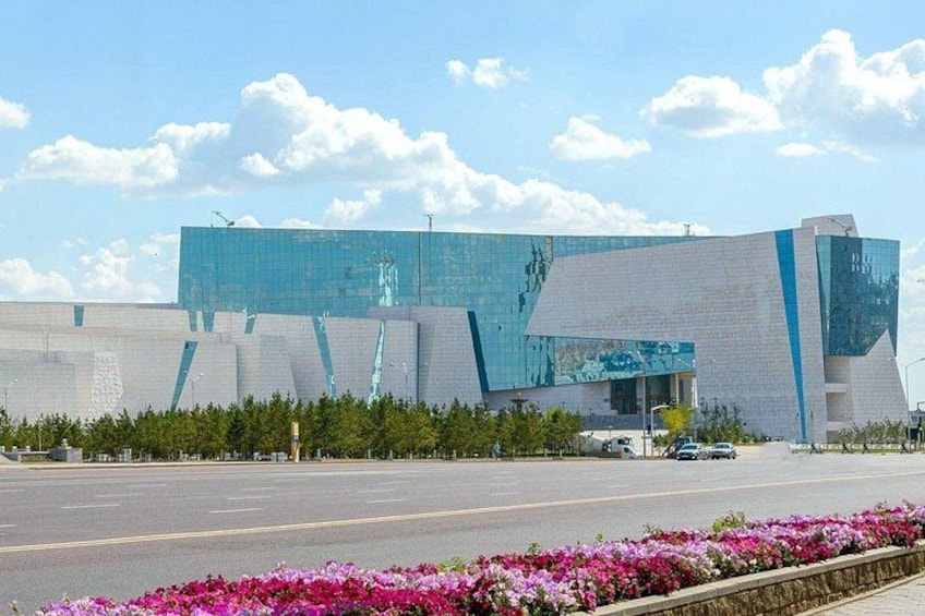 A modern building with a reflective glass facade, a wide road in front, and a flower bed with pink flowers. 2