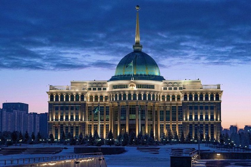 A large, historic building with a prominent dome and a tall spire, surrounded by a snow-covered plaza and modern buildings in the background. 5