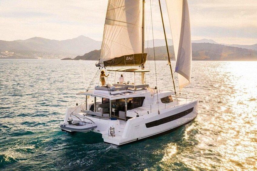 Un velero blanco con cabina y un bote auxiliar más pequeño, navegando sobre aguas tranquilas con un paisaje montañoso al fondo. 2