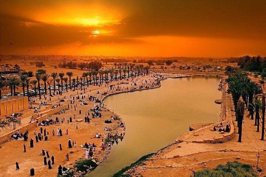 A river with people sitting and walking along the banks, palm trees lining the area, and a sunset in the background. 2