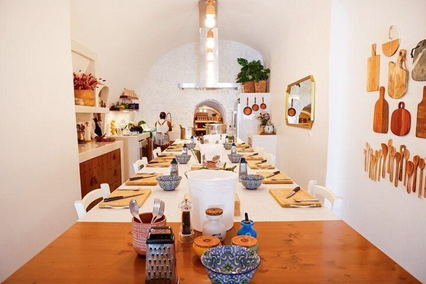 A dining area with a long table set for a meal, kitchen utensils on the wall, and a person in the background. 1