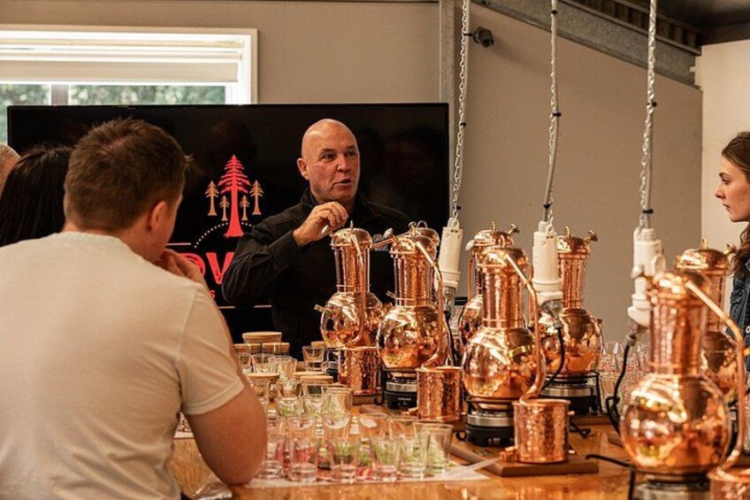 A person standing in front of a television screen with a logo, a table with copper vessels, and a person sitting at the table. 2