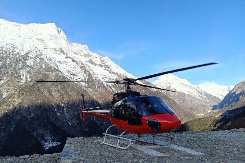 A red helicopter parked on a rocky surface with snow-capped mountains in the background. 1