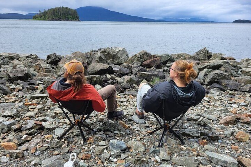 To personer sidder på stole med udsigt til en stenet kystlinje, vand og en fjer island. 1