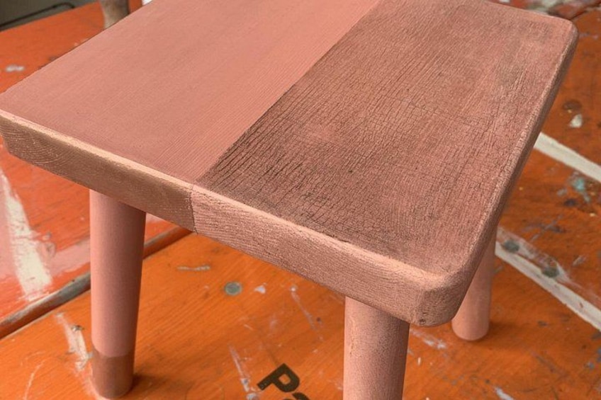 A wooden chair with a pinkish paint finish, placed on an orange surface with visible paint splatters. 2