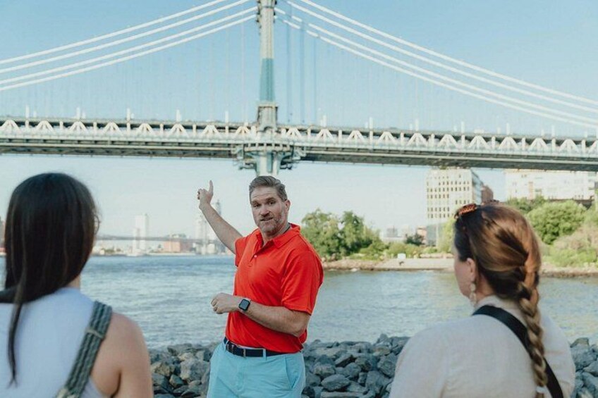 A man in a red shirt gesturing, a bridge in the background, and a river. 1