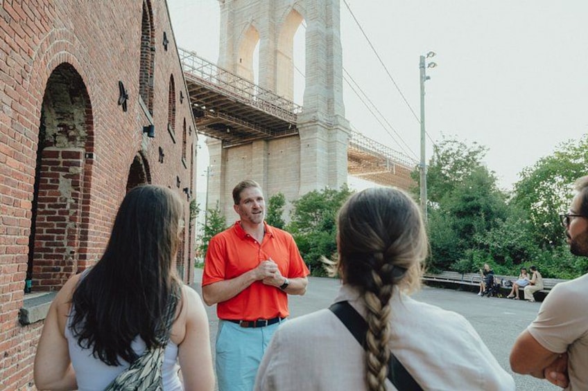 A group of people standing in front of a brick building with arched windows and a large tower in the background. 3
