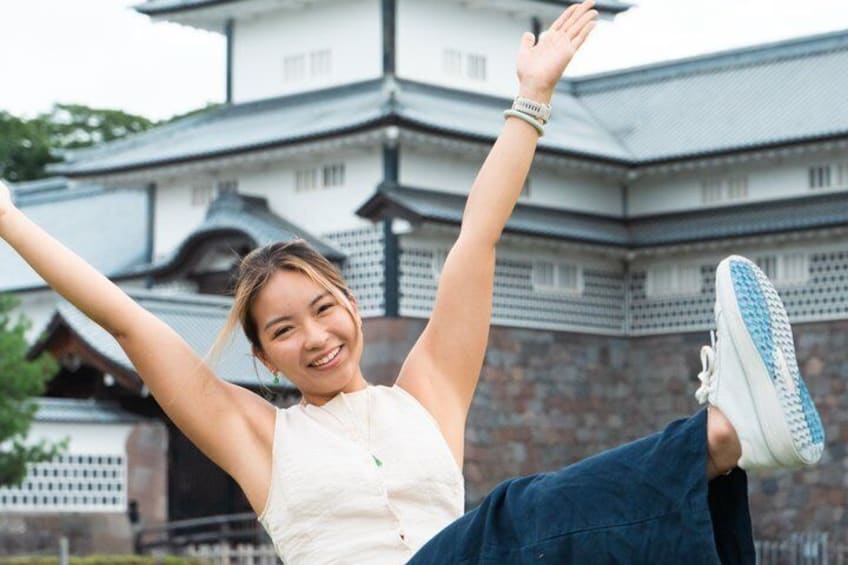 A person in a white sleeveless top and blue jeans, raising their arms in front of a traditional Japanese castle. 3