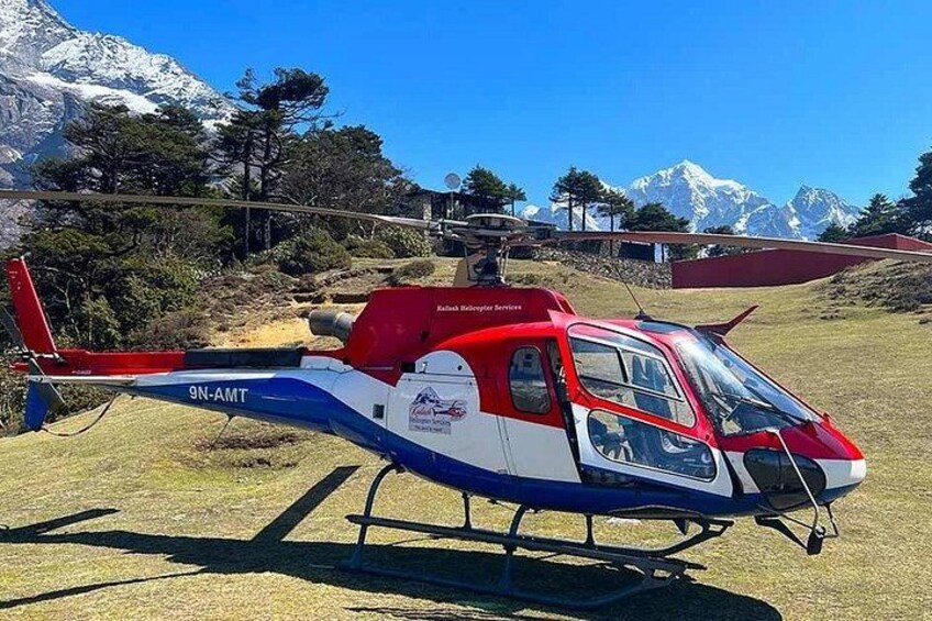A red and white helicopter with the registration number 9N-AMT parked on a grassy area with mountains in the background. 3