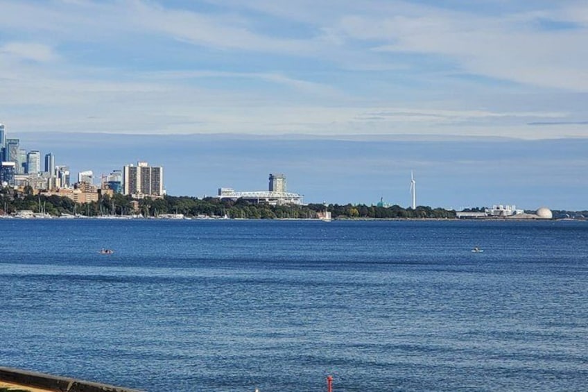A coastal cityscape with a clear sky, a large body of water, and several buildings including a wind turbine. 2