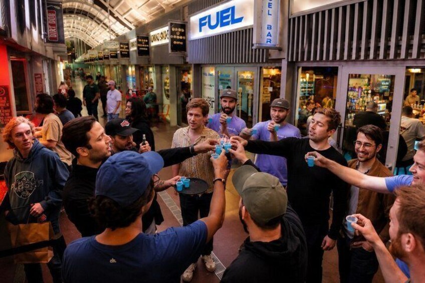 A group of people toasting with blue cups in a busy indoor setting with a visible 'FUEL' sign. 3