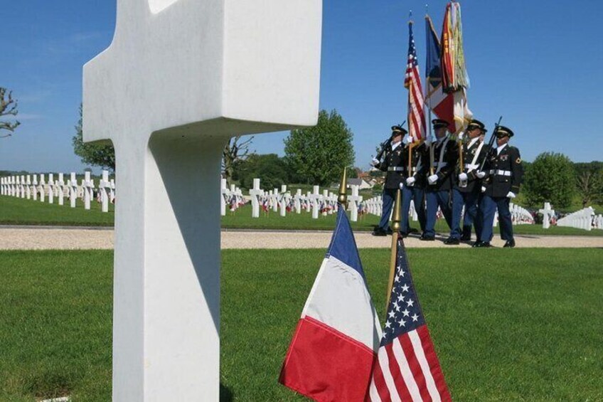 A memorial with white crosses, American and French flags, and a military ceremony in the background. 1