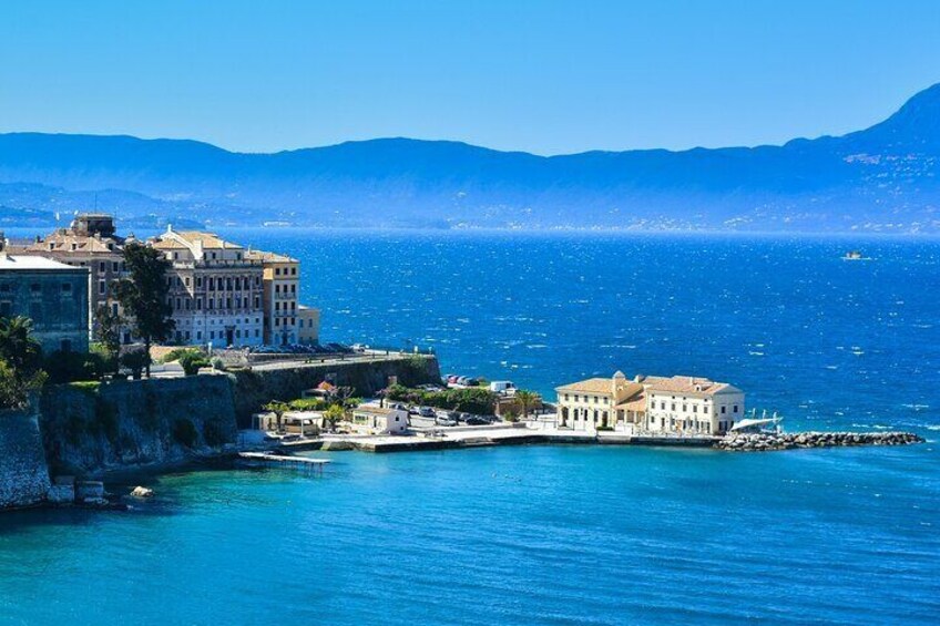 Un pueblo costero con edificios cerca del agua, un mar azul cristalino y montañas a lo lejos. 5