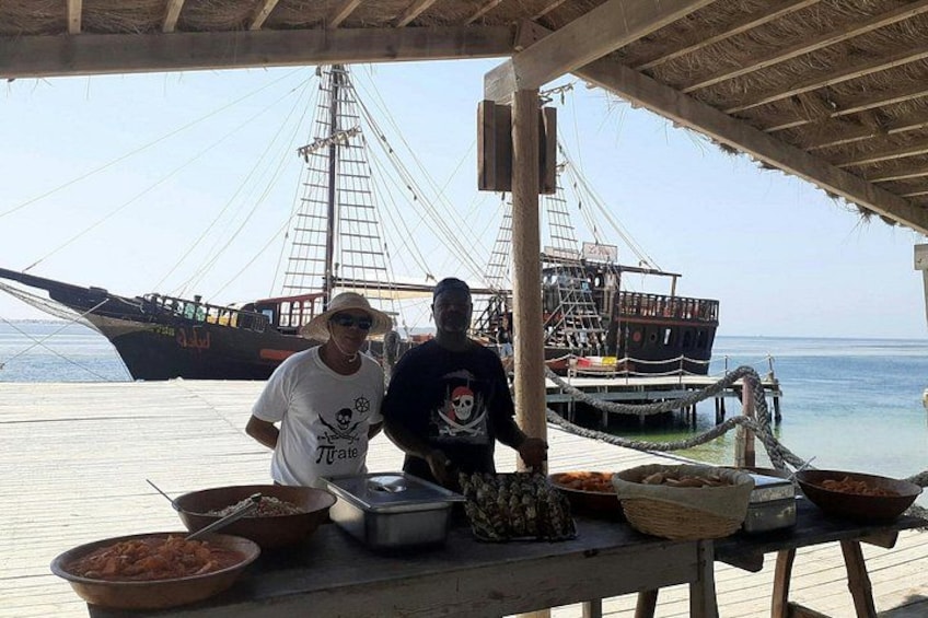Two people preparing food on a wooden dock with a large ship in the background. 4