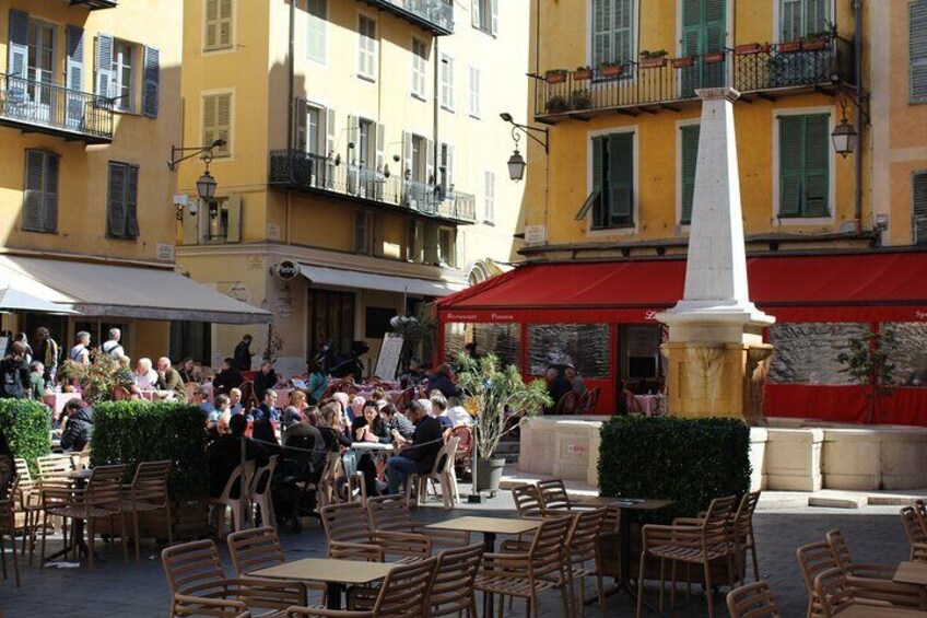 Outdoor seating at a café with a white obelisk in the center, surrounded by yellow buildings with balconies. 3