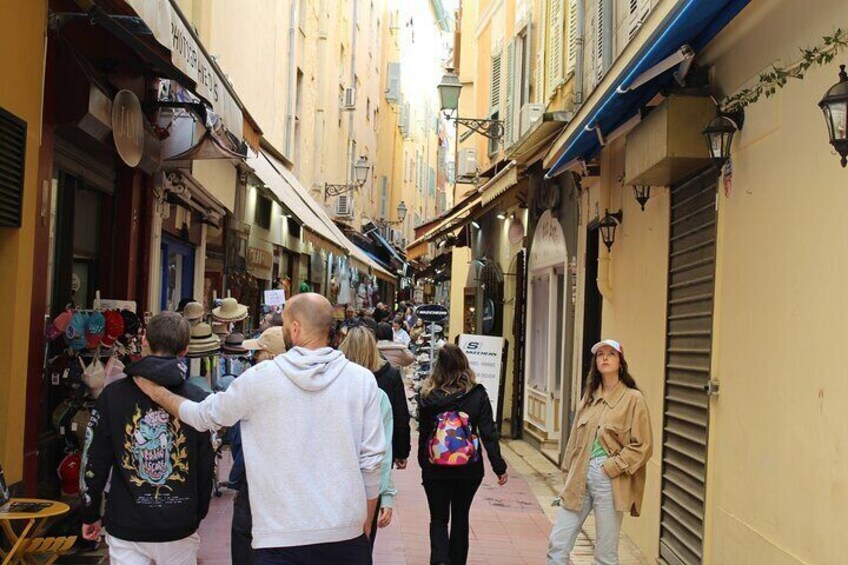 A narrow street with shops on both sides, pedestrians walking, and a person pointing. 2