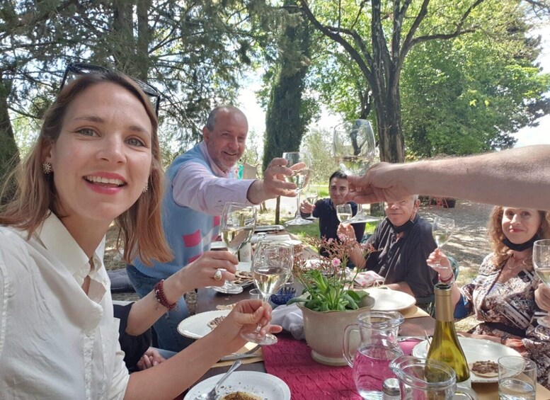 Een groep mensen proost met wijn glazen aan een buiteneet tafel, omringd door groen. 1