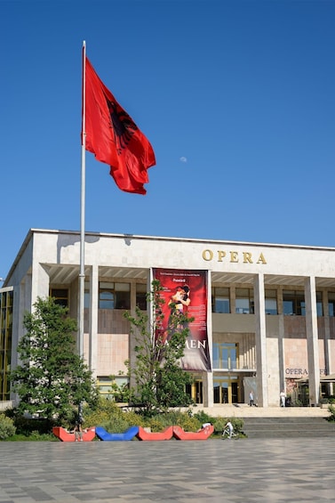 The exterior of an opera house with a red flag, a large banner, and a clear blue sky. 1
