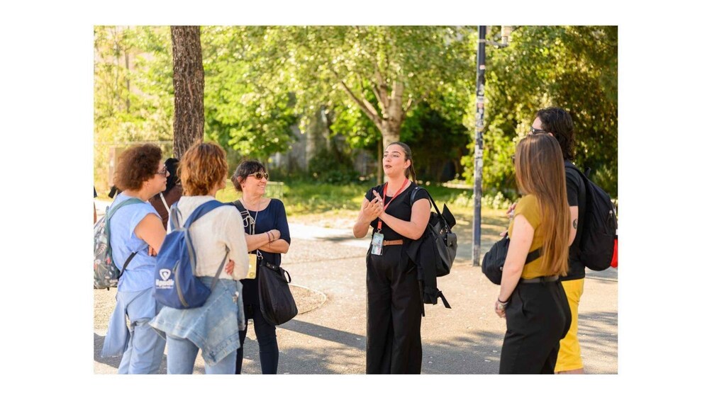 A group of people standing outdoors, engaged in conversation, with trees and a pathway in the background. 3