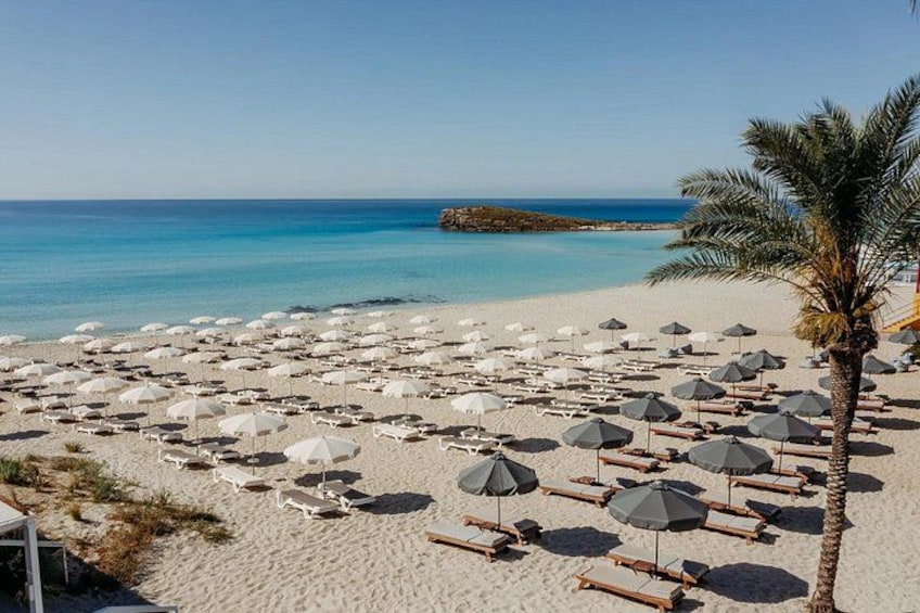 Een strand met vele ligbedden en parasols, een palmboom en een heldere blauwe zee. 2
