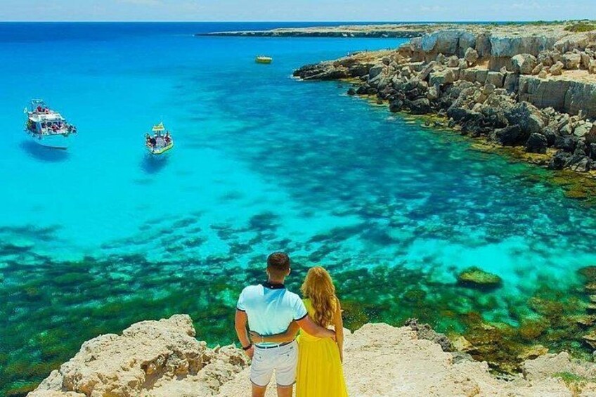 A couple overlooking a turquoise sea with boats, rocky coastline, and clear water. 1