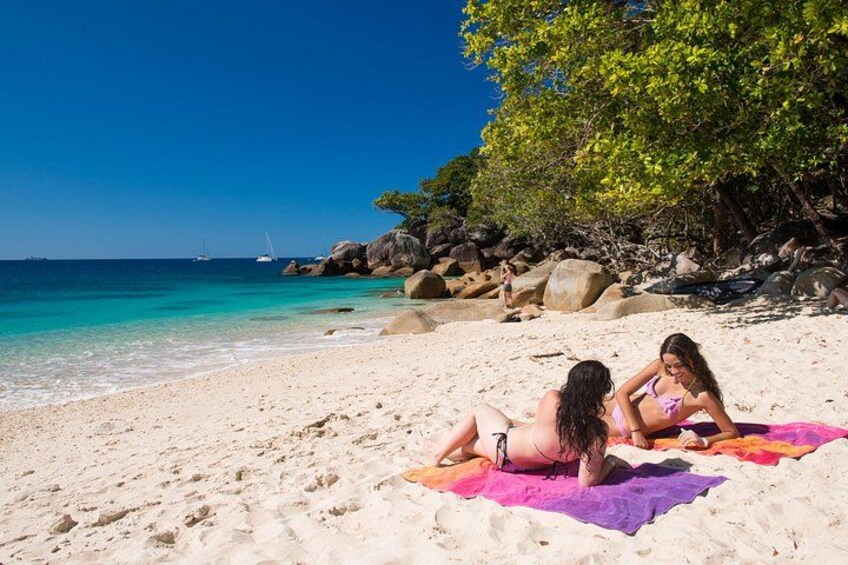 Une plage où se trouvent deux personnes sur un grand drap de bain violet, avec une eau turquoise et limpide, et des affleurements rocheux. 2