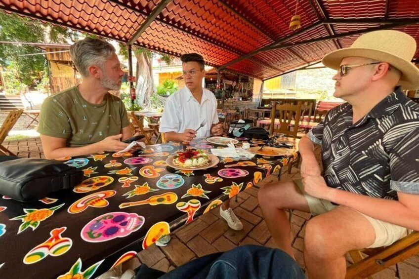 Trois personnes sont assises à une table à l’extérieur, recouverte d’une nappe colorée, sous un toit orné de tuiles rouges. 2