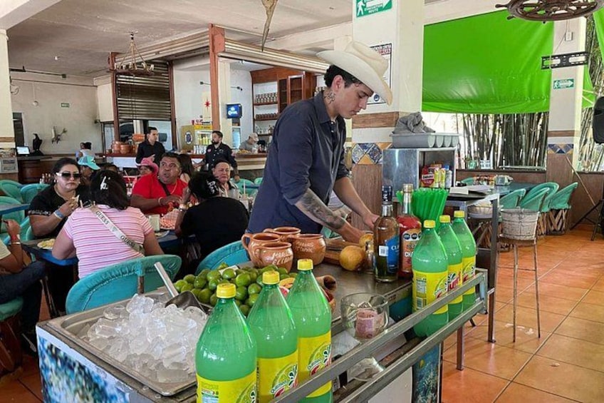 A restaurant with patrons seated at tables, a bartender preparing drinks, and a green screen in the background. 2