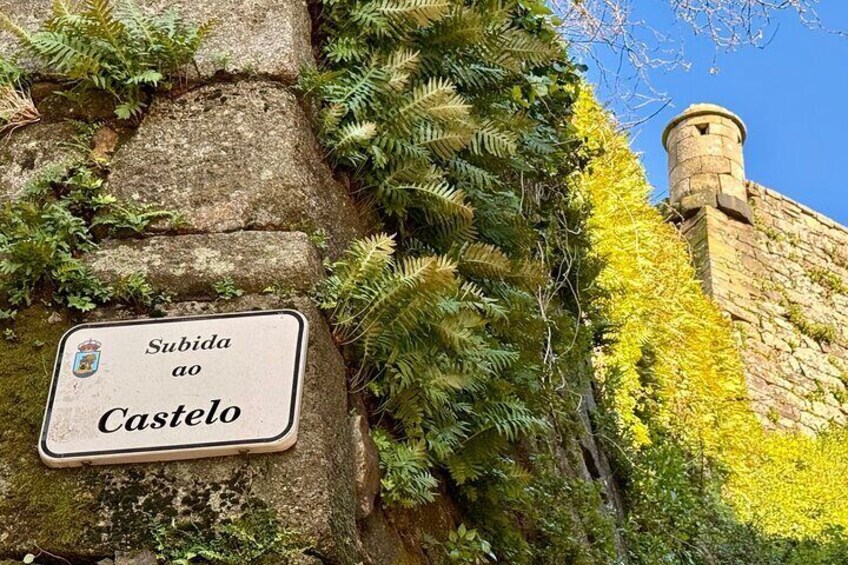 Eine Steinmauer mit einem Schild, auf dem "Subida ao Castelo" steht, und ein Turm im Hintergrund. 5