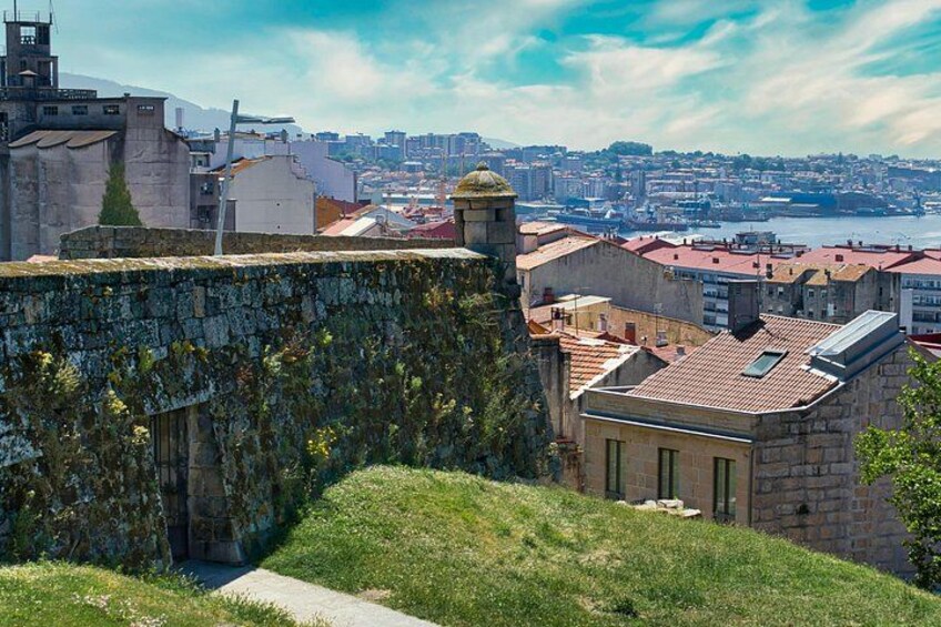 Un mur de pierre recouvert de végétation, un bâtiment avec un toit rouge et une vue sur la ville avec une étendue d’eau en arrière-plan. 3