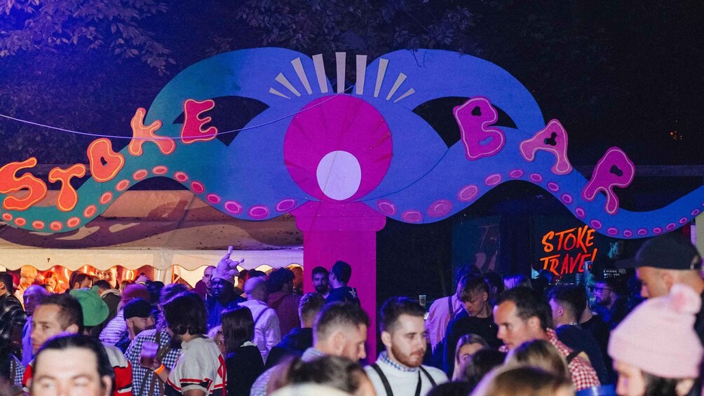 A crowd at a festival with a large tent and a sign reading "STOKE BAR". 3
