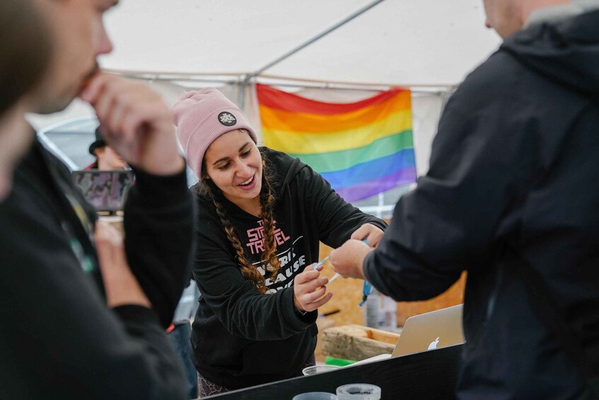 A person wearing a black hoodie with a graphic design, holding a pen, and standing at a table with a laptop. A rainbow flag is visible in the background. 4