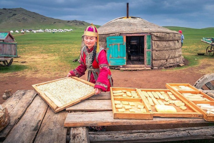 Een persoon in traditionele kledij die eten serveert bij een houten tafel buiten, met joerten en een grasachtig landschap op de achtergrond. 1