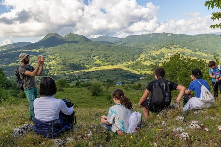 A group of people sitting and standing on a grassy hill overlooking a valley with mountains in the distance. 5