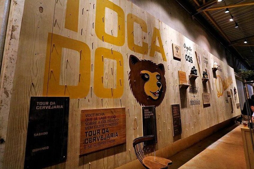 Un mur en bois orné de grandes lettres formant l’inscription « LOCA DO LOCA » et d’un visage de lion. On y trouve également des panneaux indiquant « TOUR DA CERVEJARIA » et « VOCE ACHA », accompagnés d’un texte supplémentaire. 4