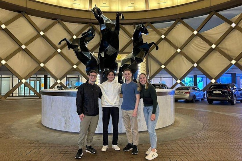 A group of people posing in front of a large sculpture of a horse in an indoor setting with a circular design and cars in the background. 3