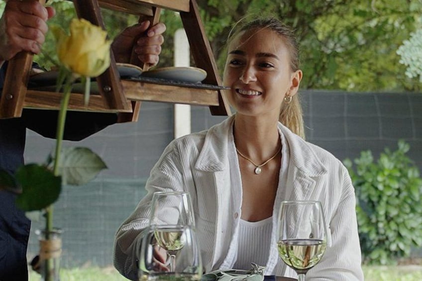 A person sitting at a table with a glass of white wine, a rose, and a wooden structure in the background. 1