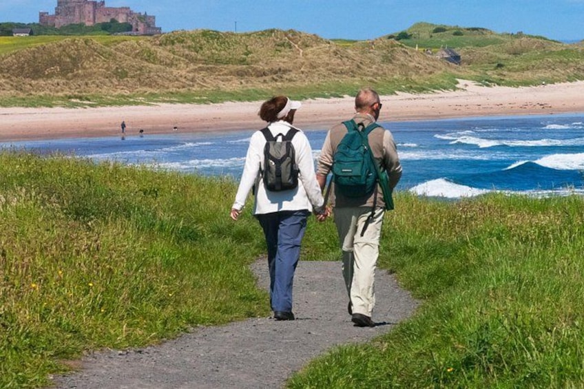 Two people walking on a path beside a beach with waves crashing onto the shore and a castle in the background. 2