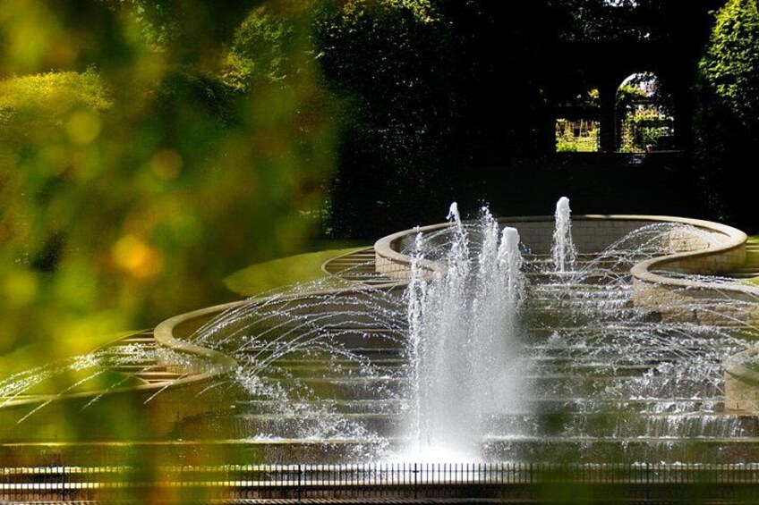 A fountain with multiple water jets in a circular basin, surrounded by greenery. 2