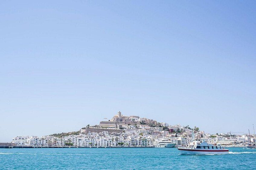 A coastal city with a prominent hilltop fortress, a clear blue sky, and a boat on the water. 1