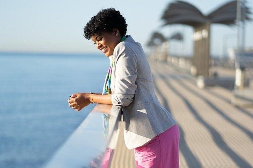 A person leaning on a railing by the water, wearing a light jacket and pink pants. 5
