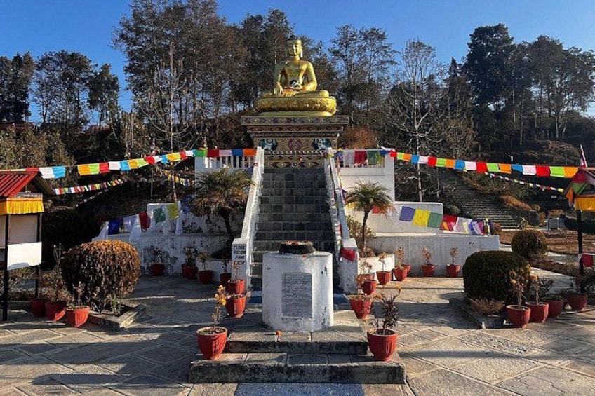 A golden statue of a seated figure, surrounded by a stone staircase, potted plants, and colorful prayer flags. 2