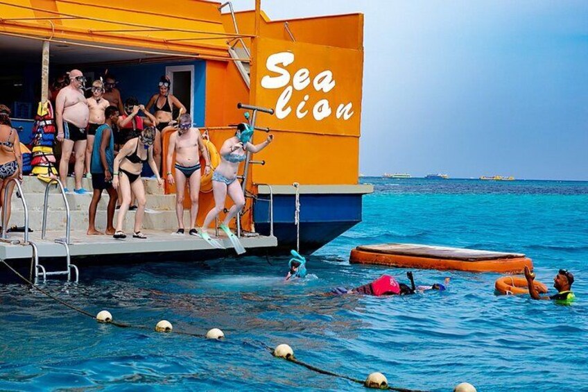 En swimmingpool med folk i badkläder, ett livräddartorn och en båt i bakgrunden. 5
