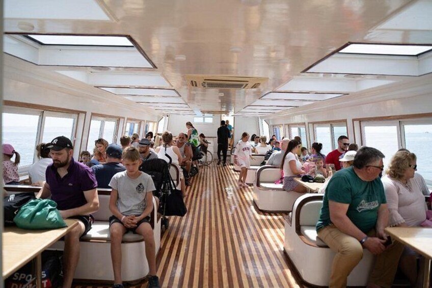 A cruise ship interior with passengers seated at tables, windows along the sides, and a central aisle. 2
