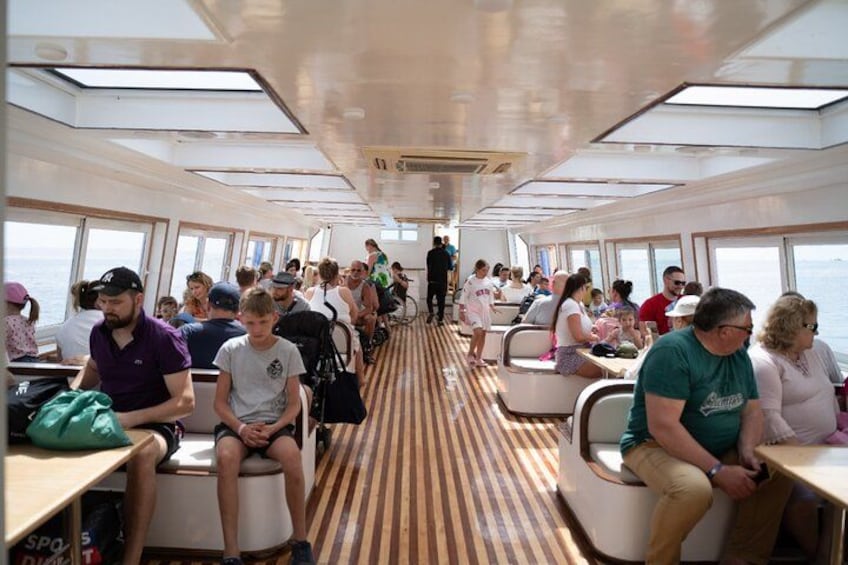 A cruise ship interior with passengers seated at tables, a central aisle, and large windows. 2