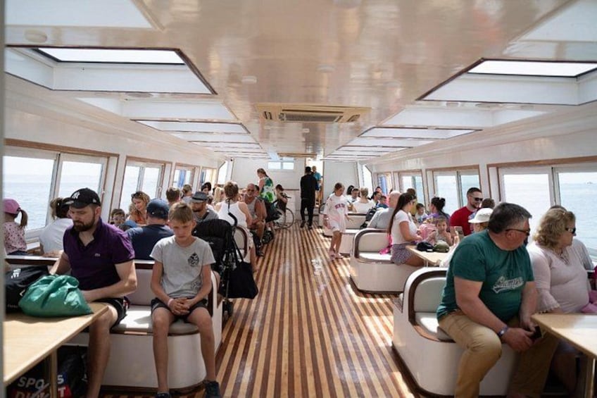A cruise ship interior with passengers seated at tables, windows along the sides, and a wooden floor. 2