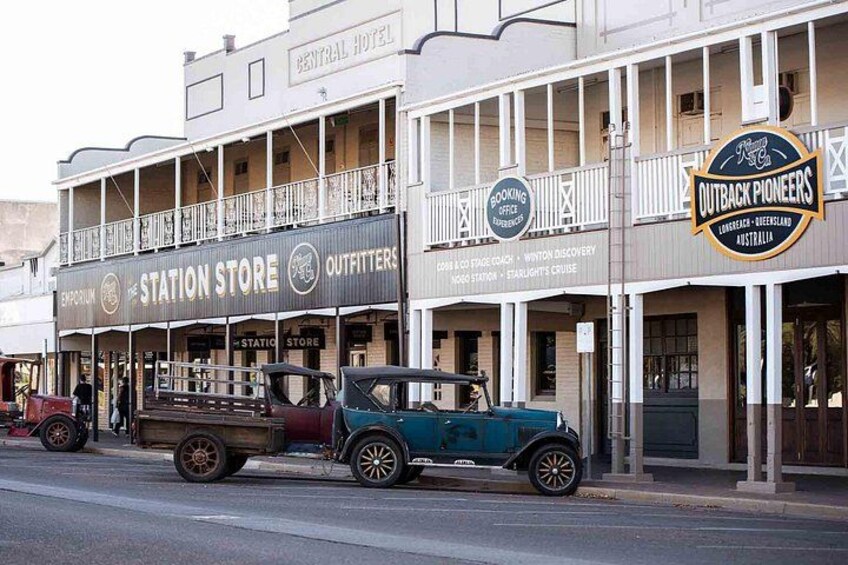 A street with vintage cars parked in front of a station store and an Outback Pioneers restaurant. 2