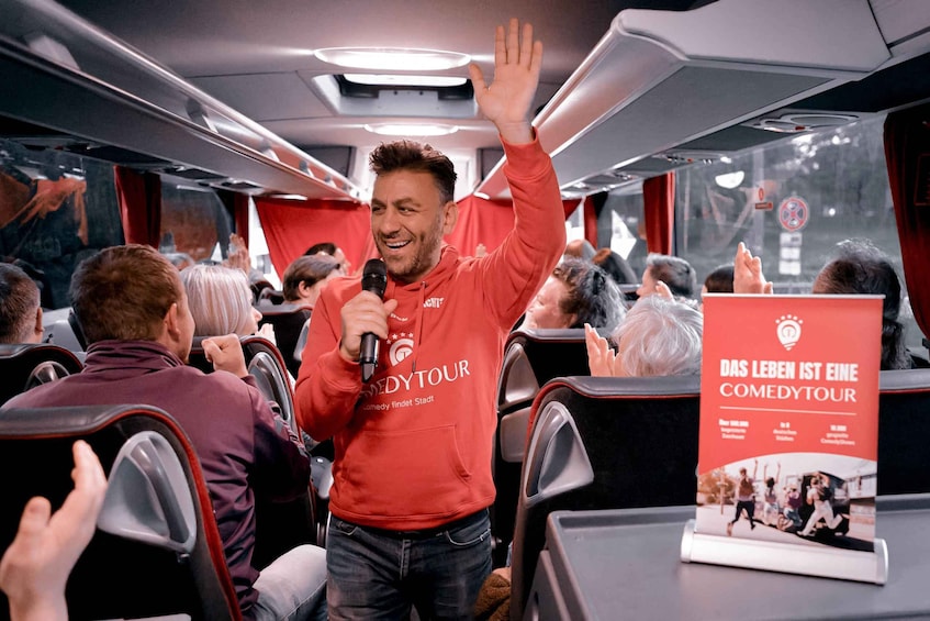 A person in a red hoodie with a microphone, standing and waving to a crowd inside a vehicle. 1