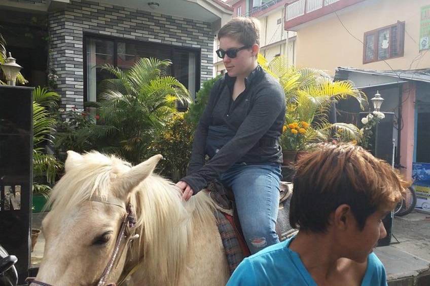 En person rider på en hest i et udendørs område med bygninger og vegetation i baggrunden. 1