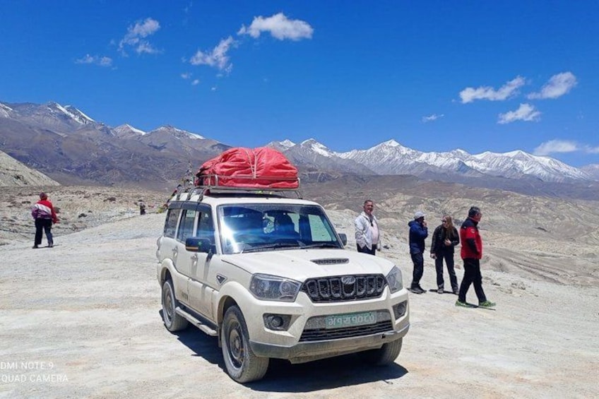 A white SUV with a red cargo bag parked on a dirt road, surrounded by mountains and a clear sky. 5